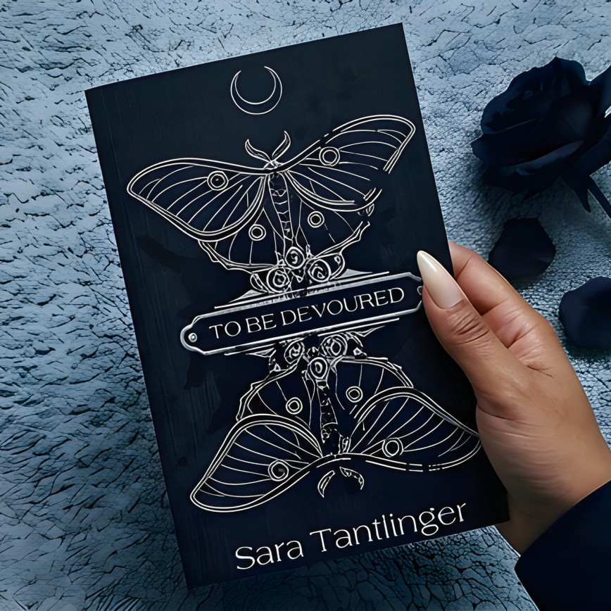 A hand with warm medium-brown skin and long almond-shaped nails holds a dark gothic paperback featuring a silver luna moth illustration and the title "To Be Devoured" against a textured grey carpet, with a black rose resting beside it.